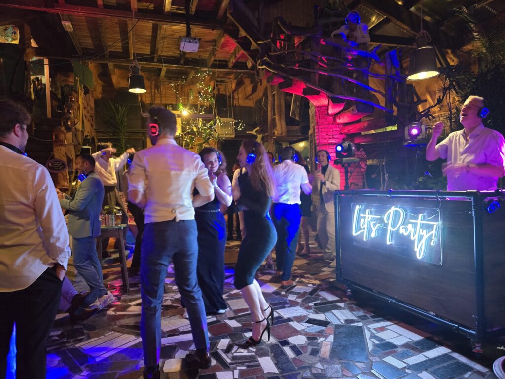 Gasten dansen met koptelefoons tijdens een silent disco, naast de industriële DJ-booth met Let’s Party-neonbord
