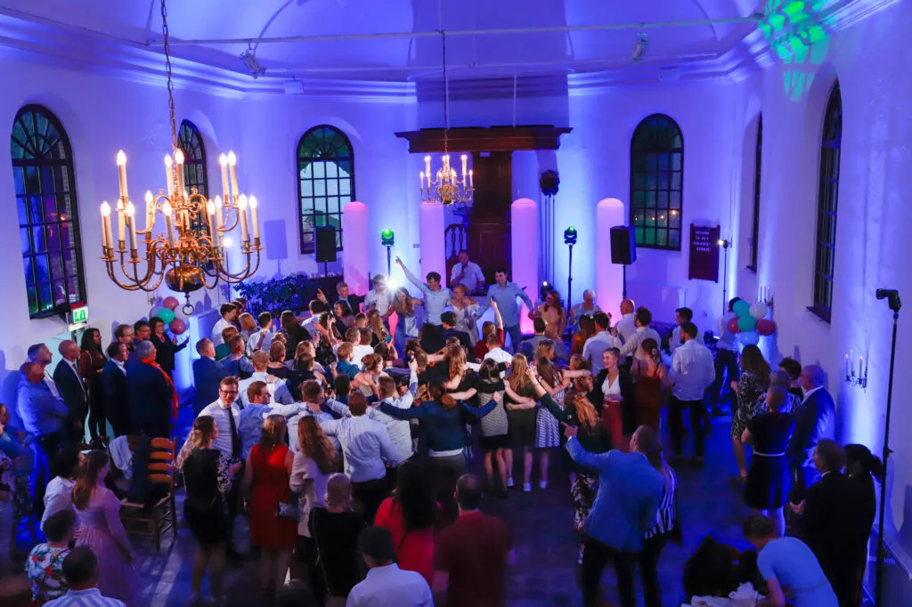 Volle dansvloer in een wit kerkje, sfeervol verlicht met blauw en paars licht en een kroonluchter