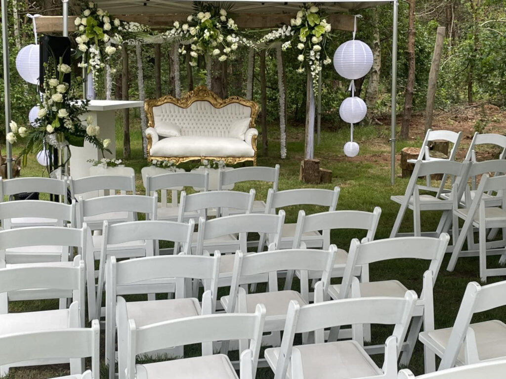 Ceremonie-opstelling tussen de bomen van Landgoed Lindehof, met witte stoelen, lampionnen en een bloemenboog
