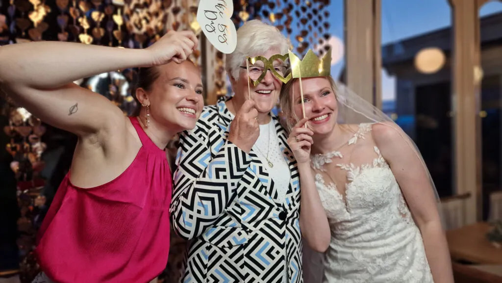Bruid, oma en een vriendin poseren lachend met props bij de photobooth in De Paalkoepel