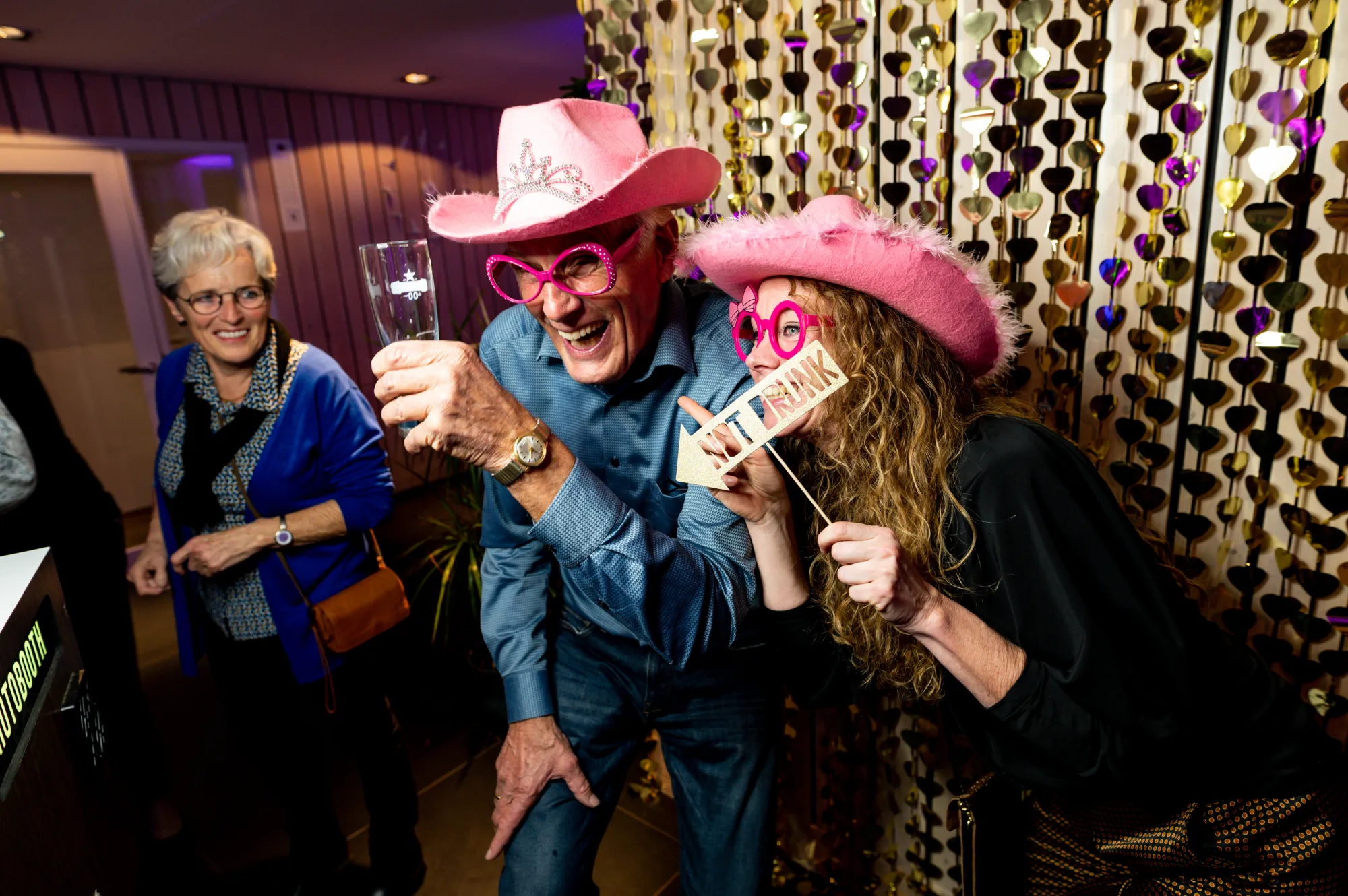 Man en vrouw met roze cowboyhoeden en feestbrillen lachen bij het gouden glittergordijn van de photobooth