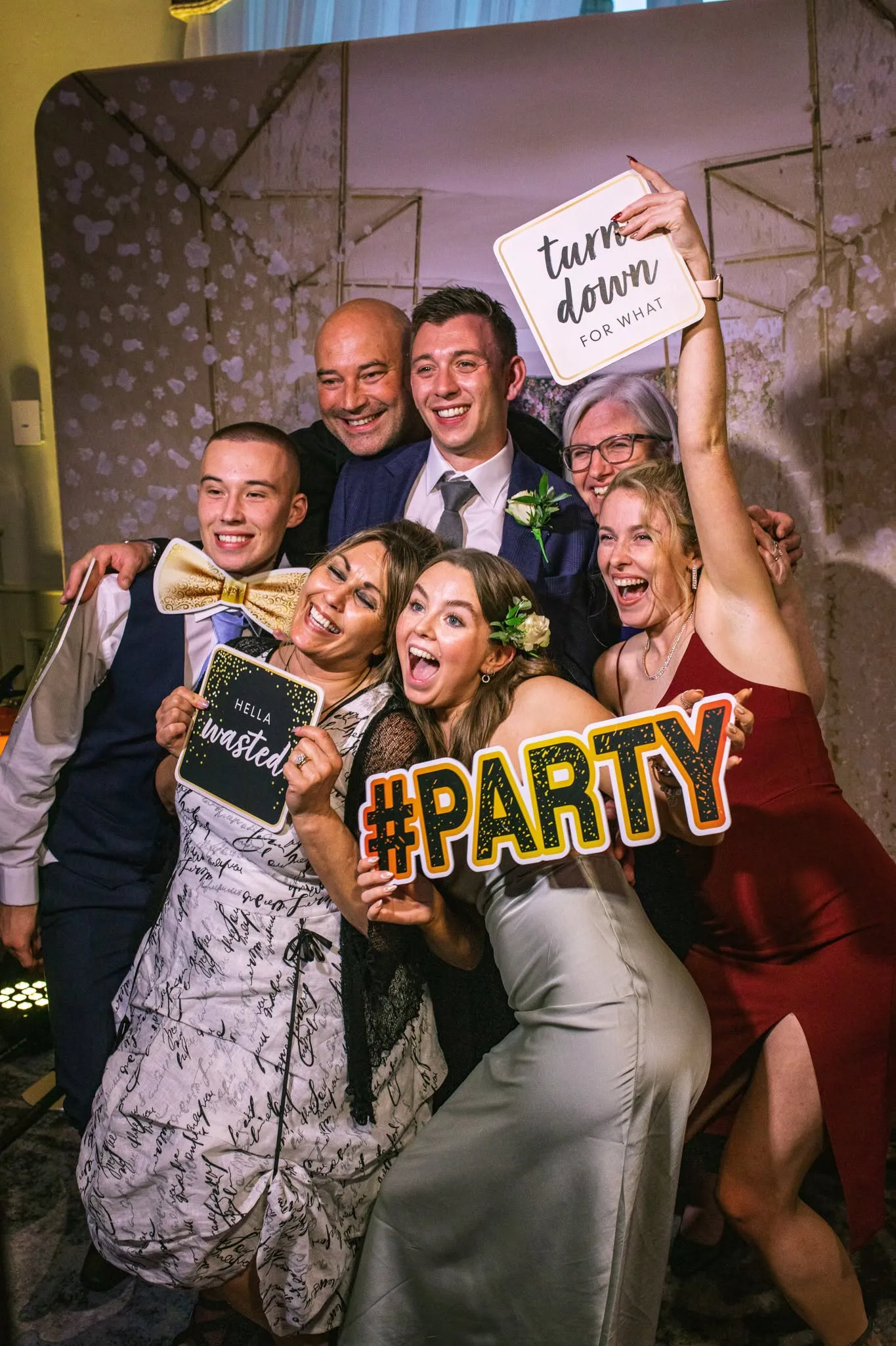 Zeven bruiloftsgasten poseren lachend in de photobooth met props en een #PARTY-bord