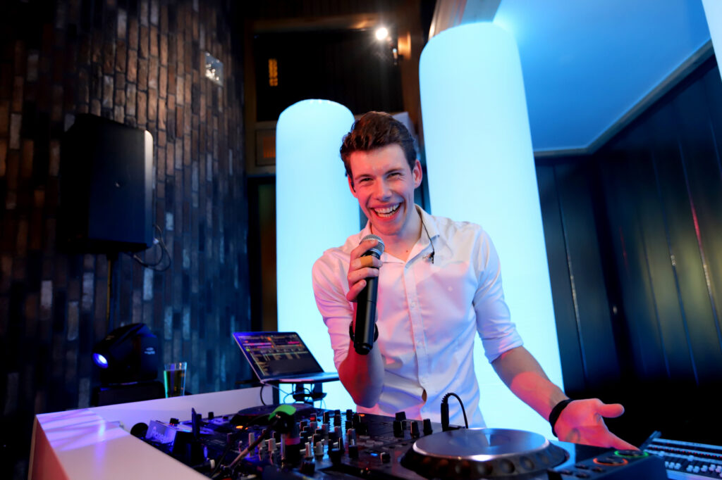 DJ Lars lacht met de microfoon in de hand achter de draaitafels, voor verlichte witte zuilen