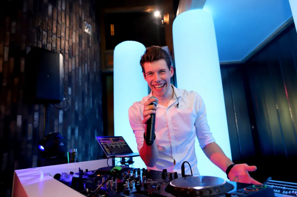 DJ Lars lacht met de microfoon in de hand achter de draaitafels, voor verlichte witte zuilen