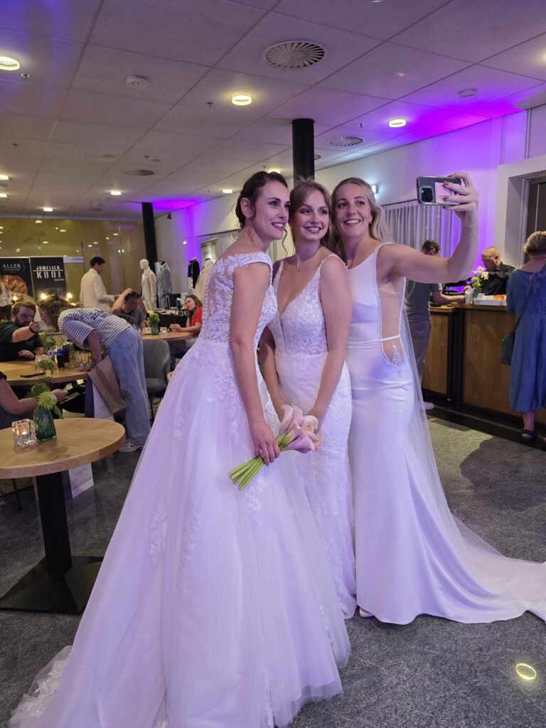 Drie bruiden in trouwjurk maken lachend een selfie op de trouwbeurs